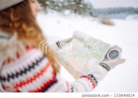 Curly-haired young woman standing in the wild with a map and compass in a snowy forest. 133649024