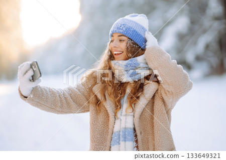 Young woman in a bright hat and scarf with a phone in a snowy forest. Travel, vacation concept. 133649321