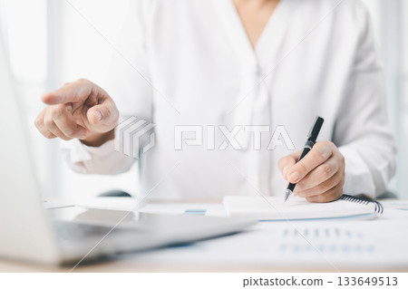 Concept of business planning and financial analysis. A businesswoman writes notes on documents with charts and reports, using laptop and calculator for budget management at modern office desk. 133649513