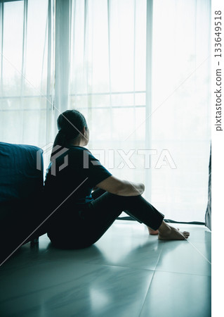 A lonely woman sits on the floor by window looking outside in dark room. Concept of depression, anxiety, sadness, loneliness, mental health problem, emotional struggle and hope for better life. 133649518