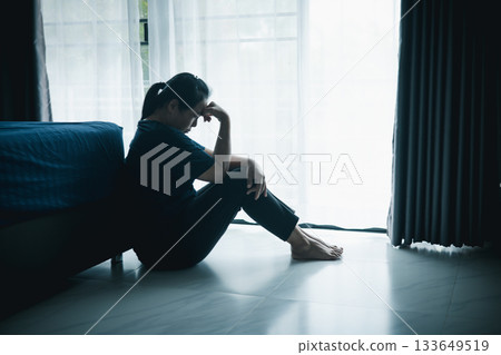 A lonely woman sits on the floor by window with hand on head in dark room. Concept of depression, sadness, anxiety, stress, mental health problem, loneliness and emotional struggle in life. 133649519