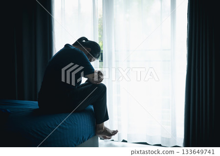 A stressed woman sitting alone on bed in dark room, holding her head down, symbolizing depression, anxiety, hopelessness, loneliness, mental health struggle, burnout and emotional pain. 133649741
