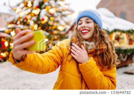 Portrait of woman with red lips taking selfie on phone while walking along Christmas streets of city Portrait of woman with red lips taking selfie on phone while walking along Christmas streets of city 133649782