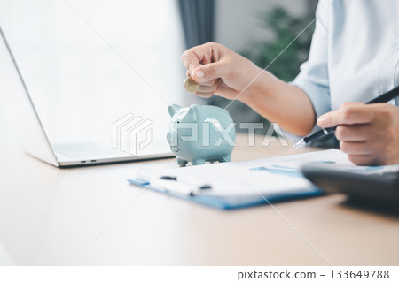 Concept of saving money and financial planning. A businesswoman puts coin into blue piggy bank on office desk with documents and laptop, symbolizing investment, budget management and future growth. 133649788
