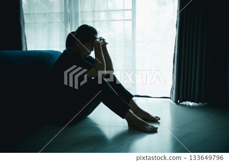 A lonely woman sits on the floor by window with hand on head in dark room. Concept of depression, sadness, anxiety, stress, mental health problem, loneliness and emotional struggle in life. 133649796