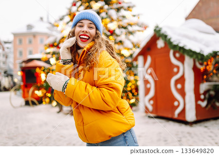 A woman in a bright jacket and red lips walks through a Christmas market and drinks a hot drink. 133649820