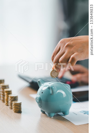 Concept of saving money and financial planning. Businesswoman puts coin into blue piggy bank on office desk with documents and laptop, symbolizing investment, budget management and future growth. Concept of saving money and financial planning. Businesswoman puts coin into blue piggy bank on office desk with documents and laptop, symbolizing investment, budget management and future growth. 133649833