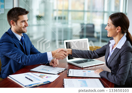 Two business people shaking hands across a table in a modern office environment, with documents, charts, and a laptop visible. Two business people shaking hands across a table in a modern office environment, with documents, charts, and a laptop visible. 133650103
