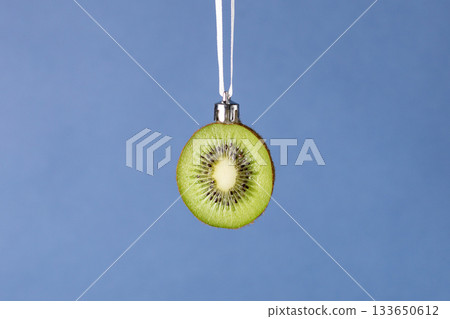 Creative christmas ornament: fresh kiwi slice hanging on white ribbon on blue background Creative christmas ornament: fresh kiwi slice hanging on white ribbon on blue background 133650612