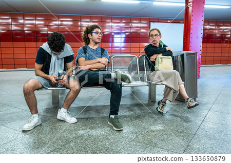 Berlin, Germany, August 10, 2023. A subway station in Frankfurt am Main, characterized by its red and orange tiled walls. On a bench, two young men and a middle-aged woman wait for the train to pass. 133650879