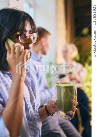 Young woman holding matcha and talking on phone in relaxed office environment Young woman holding matcha and talking on phone in relaxed office environment 133651928