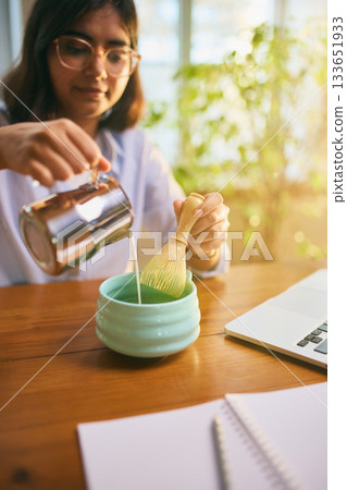 Woman pouring milk into matcha bowl while preparing beverage at desk 133651933