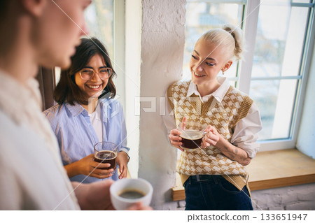 Coworkers laughing together while holding coffee near a bright window 133651947