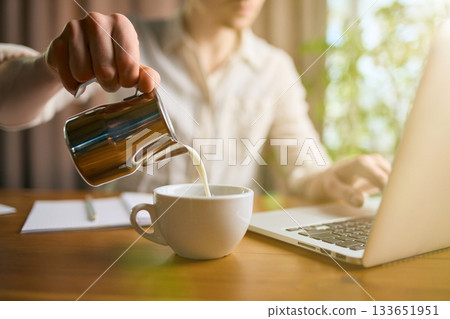 Man pouring milk into coffee cup while working on laptop Man pouring milk into coffee cup while working on laptop 133651951