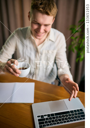 Smiling man working on laptop with coffee cup in hand at wooden desk 133651953