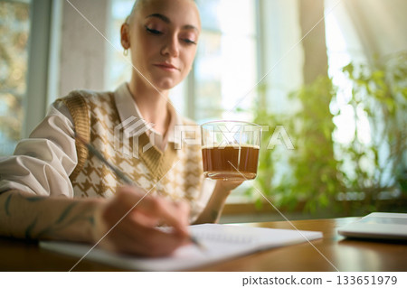 Woman holding coffee while writing notes in bright office Woman holding coffee while writing notes in bright office 133651979