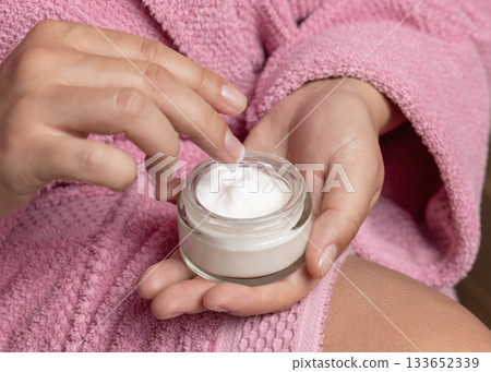 Young woman in pink bathrobe touching cream with finger and holding a cosmetic jar closeup 133652339