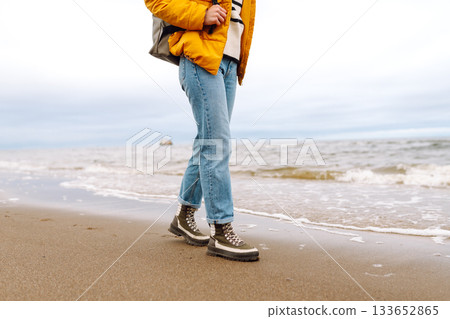 Woman traveler walks in boots along the seashore. Happy traveler with a backpack enjoys the sea air. Woman traveler walks in boots along the seashore. Happy traveler with a backpack enjoys the sea air. 133652865