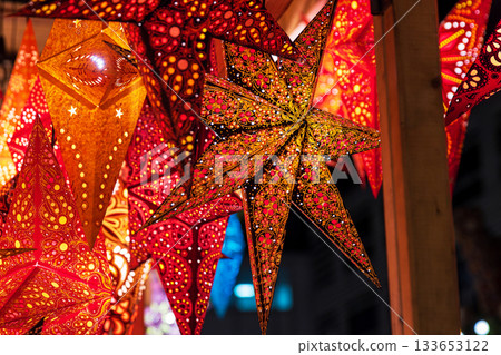 Vibrant star shaped lanterns in red, orange, gold patterns glowing hanging at a festive christmas market stall. Detailed perforated designs create sparkling light effects, Vibrant star shaped lanterns in red, orange, gold patterns glowing hanging at a festive christmas market stall. Detailed perforated designs create sparkling light effects, 133653122
