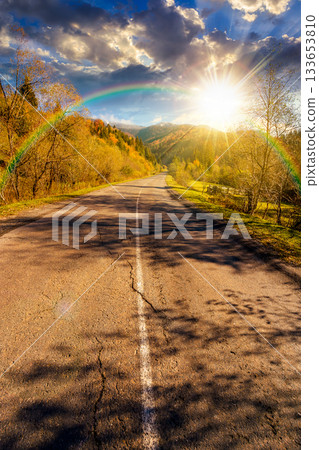 road through mountain landscape in autumn at sunset. forest in fall colors under blue sky and rainbow in evening light. snow covered peak in the distance. scenic view of rural valley road through mountain landscape in autumn at sunset. forest in fall colors under blue sky and rainbow in evening light. snow covered peak in the distance. scenic view of rural valley 133653810