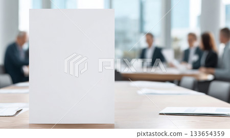 Large blank table tent mockup positioned centrally on a conference table, surrounded by blurred professionals engaged in discussion, ideal for presentations 133654359