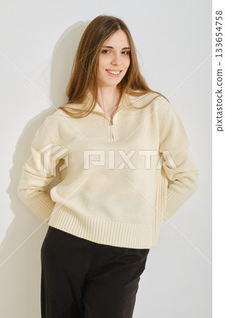 A young woman poses in a stylish, cream-colored sweater paired with black pants. A young woman poses in a stylish, cream-colored sweater paired with black pants. 133654758