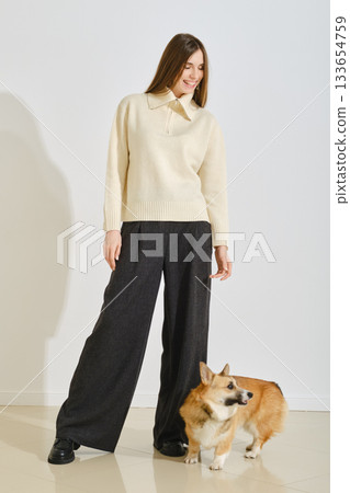 Playful woman in ivory sweater and loose pants playing with her dog in bright room 133654759