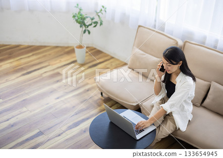 一位面帶微笑的年輕女子坐在客廳沙發上,一邊使用筆記型電腦工作,一邊用智慧型手機打電話。 一位面帶微笑的年輕女子坐在客廳沙發上,一邊使用筆記型電腦工作,一邊用智慧型手機打電話。 133654945