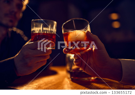 Two hands raising whiskey glasses for evening toast Two hands raising whiskey glasses for evening toast 133655172