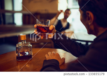 Man raising whiskey glass toward man in background. Man raising whiskey glass toward man in background. 133655179