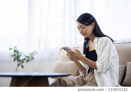 一位面帶微笑的女士一邊在沙發上放鬆，一邊使用智慧型手機。 133655181