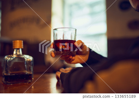 Hand holding whiskey glass beside bottle on wooden table. 133655189