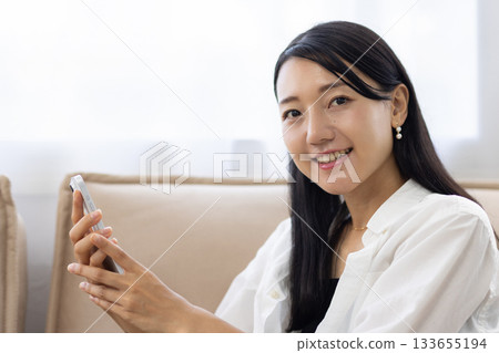 一位面帶微笑的女士一邊在沙發上放鬆，一邊使用智慧型手機。 133655194