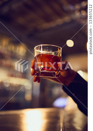 Cocktail with rich amber color held high against blurred bar interior Cocktail with rich amber color held high against blurred bar interior 133655212