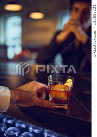 Bartender sliding amber drink toward thoughtful customer at bar Bartender sliding amber drink toward thoughtful customer at bar 133655221