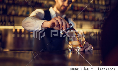 Bartender pouring spirit from jigger into iced glass on polished bar counter Bartender pouring spirit from jigger into iced glass on polished bar counter 133655225
