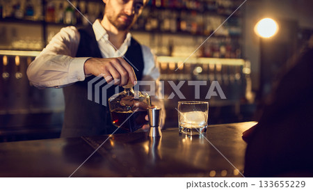Bartender pouring dark spirit from bottle into jigger beside iced glass 133655229