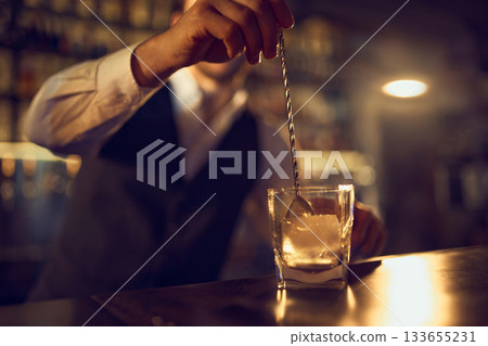 Bartender stirring large ice cube in glass with long bar spoon 133655231