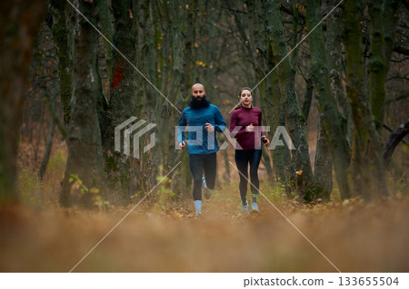 Couple running between tall forest trees on leaf-covered autumn pathway 133655504