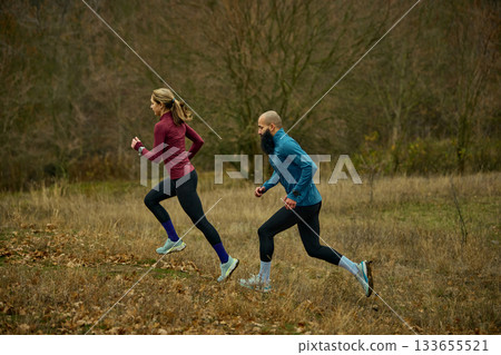 Side view of couple trail running across dry grass field in autumn woods Side view of couple trail running across dry grass field in autumn woods 133655521