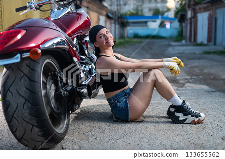 Brunette woman in denim shorts repairs a red motorcycle. Brunette woman in denim shorts repairs a red motorcycle. 133655562