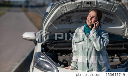 Caucasian woman laughing and calling on phone asking for help standing near car with open hood. Caucasian woman laughing and calling on phone asking for help standing near car with open hood. 133655749