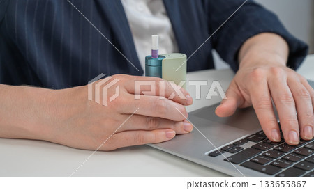 Business woman smoking an electronic cigarette while working on a laptop. Tobacco heating system. Business woman smoking an electronic cigarette while working on a laptop. Tobacco heating system. 133655867