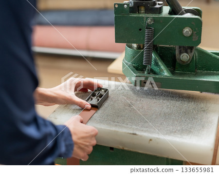 A woman makes holes in a leather belt in a workshop. 133655981