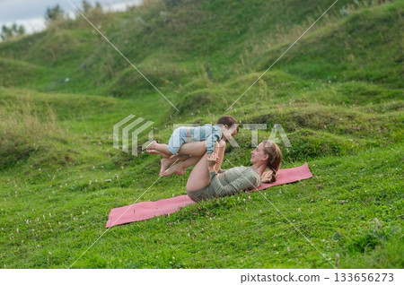 Caucasian woman doing yoga with her little son outdoors. Caucasian woman doing yoga with her little son outdoors. 133656273