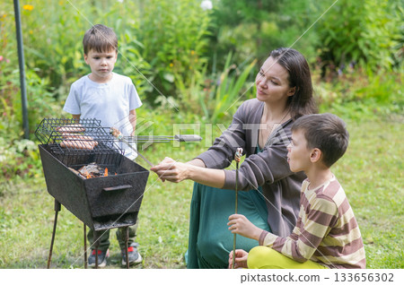 Caucasian woman grilling sausages on a barbecue with her two sons.  133656302