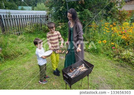 Caucasian woman grilling sausages on a barbecue with her two sons.  133656303
