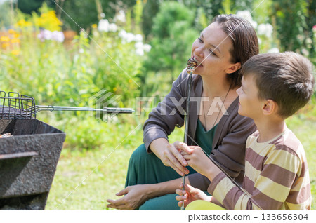 Caucasian woman grilling sausages on a barbecue with her son.  133656304