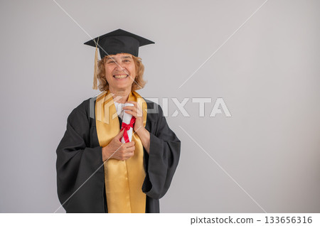 Happy senior woman in graduation gown holding diploma on white background.  133656316