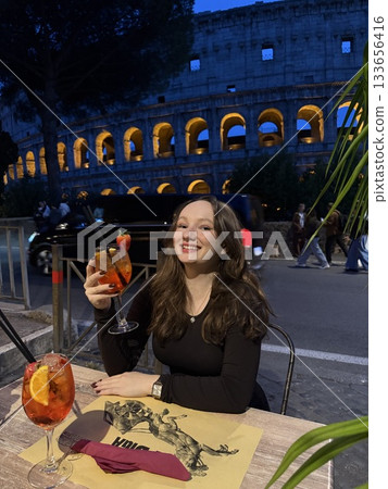 Rome, Italy 11.29.2025 Aperol in front of the Colosseum girl sitting in a restaurant near the Colosseum, eating pizza in evening, people walking in background, talking, blogging, and seeing sights. 133656416